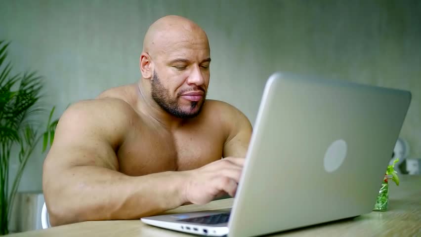 Brutal male bodybuilder working at a laptop and eating healthy salad of gre...