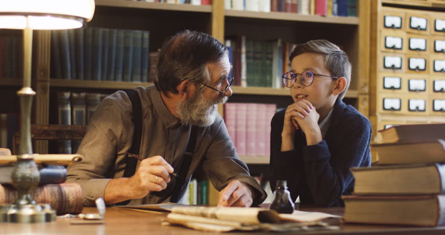 Old grandfather in glasses and with a beard writing something with an ink and talking with his grandson at the table in the library with many books.