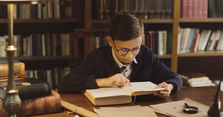 Portrait shot of the teen boy in glasses reading a book while sitting at the desk in the library, then flipping a page.