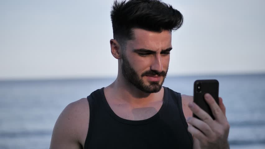 Young shirtless muscular handsome man doing videochat with smartphone by the sea at sunset