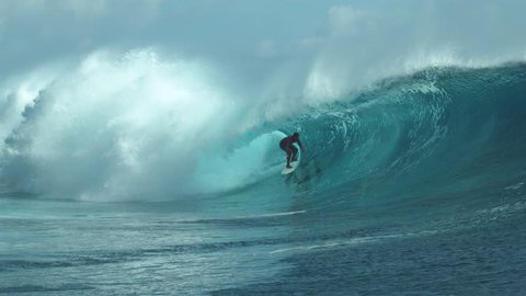SLOW MOTION: Giant emerald tube wave crashes wildly behind pro sportsman surfing near a popular surf spot in French Polynesia. Crystal clear ocean water spraying in air as barrel wave approaches coast