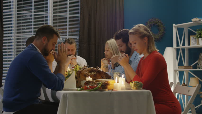 Men and women giving worship before starting delicious meal at table having Thanksgiving holiday celebration all together