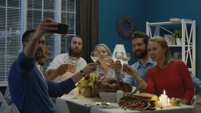 Man taking photo of diverse happy friends gathering at table on Thanksgiving day celebration and posing while having fun at home
