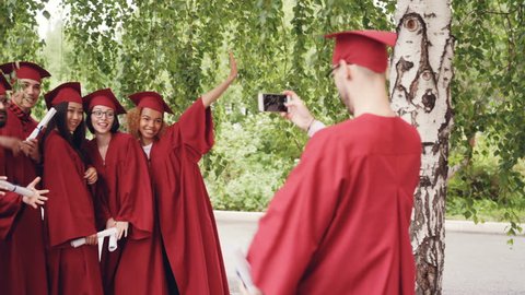 Graduating student is recording video of his friends in gowns holding diplomas, waving hands and posing looking at smartphone camera. People and technology concept.