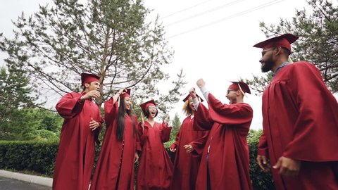 Low angle shot of joyful men and women graduating students throwing mortar-boards in the air and laughing. Beautiful summer nature and sky is visible.