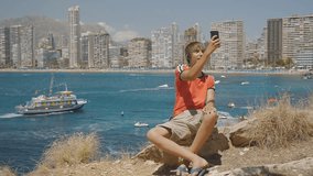 Caucasian teen boy makes video call on smartphone showing sights around on sea coast and skyscrapers skyline background