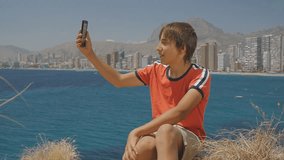 Handsome teen boy makes video call on smartphone showing sights around on sea coast city skyline background