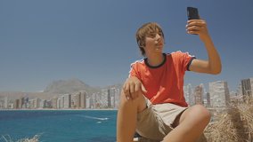 Cute teen boy makes video call on smartphone showing sights around on sea coast city skyline background