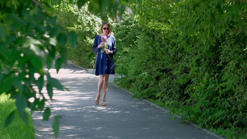 A young woman in a blue dress and sunglasses walking in the city Park. Lady walking down the alley with a pink peony flower