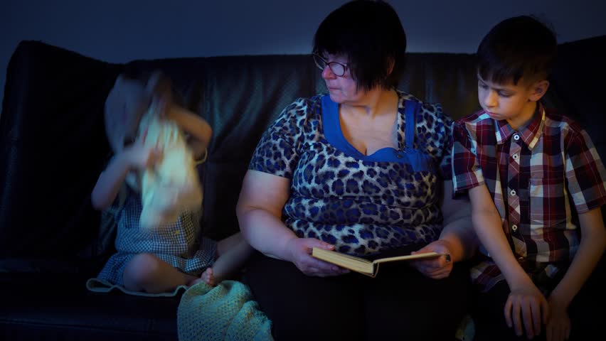 Grandmother reads a book at home to her grandson and granddaughter in the evening at home on the couch. Family vacation is a joint time for reading.