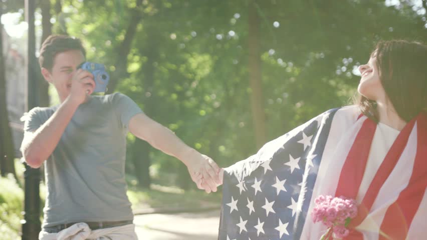 Cute portrait couple walk feel happy man take photo woman with American flag in park sunset sunlight slow motion caucasian family havingfun cheerful lovers outdoors young fun boyfriend city date