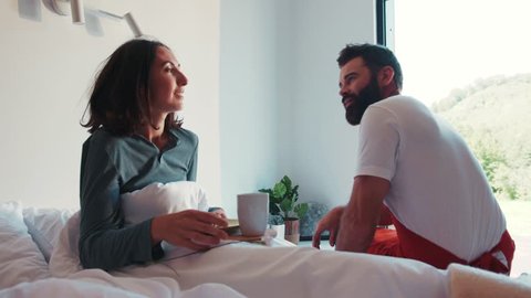 Attractive strong romantic bearded man in a red apron brings his wife a flower and breakfast in bed, talking to each other. Couple goals, true love, love story. Happy together, man’s power.
