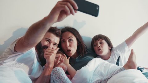 Cheerful happy family taking selfie in a bed, thumbs up, smiling. Having fun, social networks, cool parents. Cute kids. Family time, joy of life.