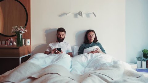 Lovely romantic couple had a fight, laying together in bed, silently using their phones. Glancing with love at each other. Bad mood, being online, modern communication. Cozy light bedroom. Love story.