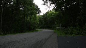 Clip of three teenage boys long boarding down a wooded road. The road has green vegetation and trees.