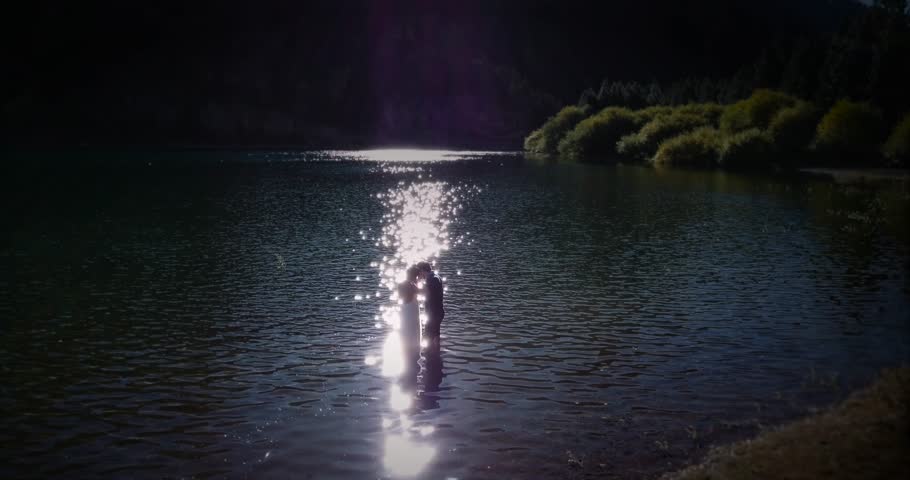 Couple of newlyweds kissing and hugging in the lake after their marriage. Silhoette of the groom and the bride inside the lake at sunset. 4k