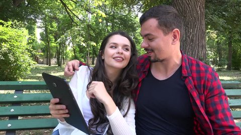 Lovely tender young family couple sit in the park bench looking tablet computer 