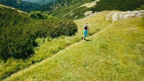 Running woman on the top of mountain. Runner is jogging in summer sunny day around trees and mountains background under sun light with blue sky, Drone 4K Video