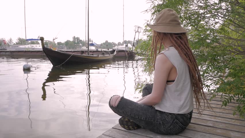 young woman traveler sitting on wooden pier near the river with boats on background	