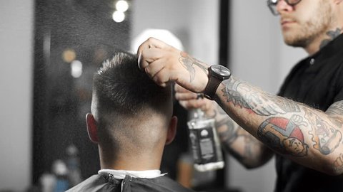Barber wets hair by spray and combs them, tattoed barber makes haircut for customer at the barber shop, man's haircut and shaving at the hairdresser, barber shop and shaving salon