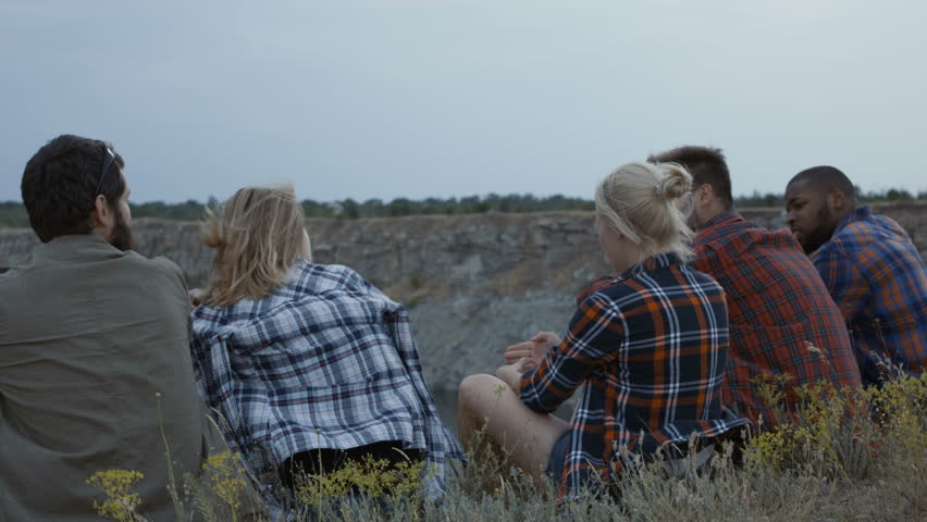 Back view of casual men and women sitting on green cliff above lake relaxing and raising hands in fun while taking selfie