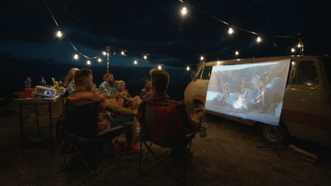 Friends gathering in campsite around bonfire and watching movie with projector on van side in dark evening