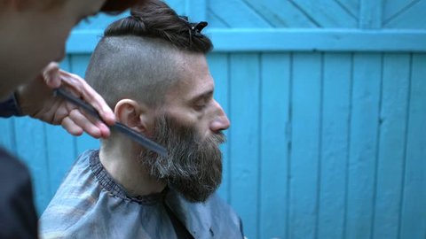 Young woman barber triming beard of  middle-aged hipster man in experimental outdoor barbershop. 
