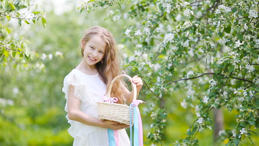 Adorable little girl with busket in blooming apple tree garden on spring day in search of eggs for Easter