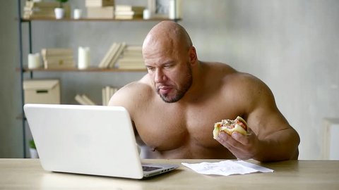 Shirtless weghtlifter holding fat junky hamburger while reading something online.