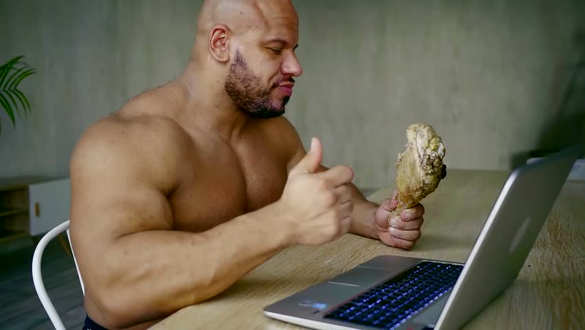 Side view shot of a giant, muscular sportsman eating cooked turkey leg at t...