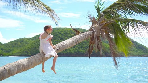 Little girl at tropical beach sitting on palm tree and having a lot of fun. Kid on caribbean vacation in Antigua island