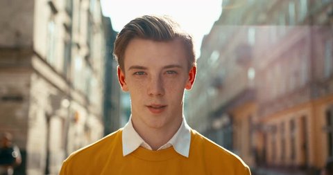 Close-up portrait of the charming young hipster with freckles looking at camera in sunbeams. 4k footage. City center location.