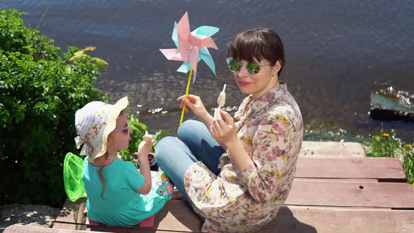 Mother and daughter sitting at wooden pier enjoying sea view on a sunny day. Young woman and little girl eating ice cream.