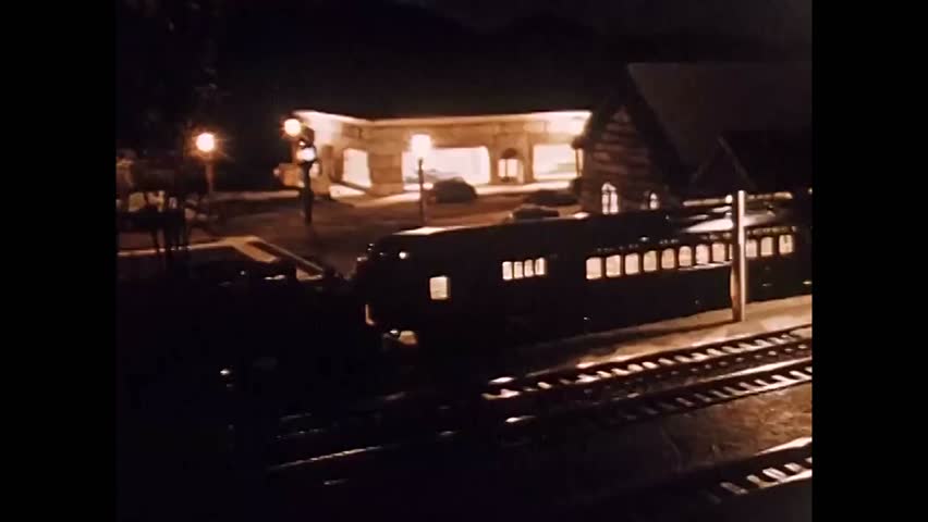 CIRCA 1950s - A model train leaves a station and travels through a city and on a bridge over a river.