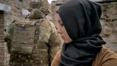 Armed soldier with weapon passing by muslim woman in hijab, woman standing in ruined abandoned building, war concept