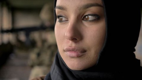 Close portrait of young confident muslim woman turning and looking at camera, armed soldier in background, war concept