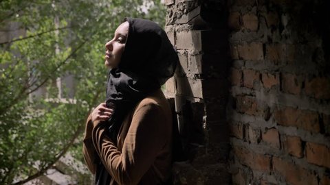 Young muslim woman in black hijab holding her hands and looking upwards, standing in abandoned brick building