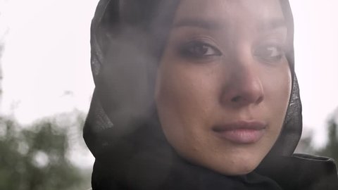 Portrait of young sad muslim woman in hijab looking at camera and crying, rainy weather in background