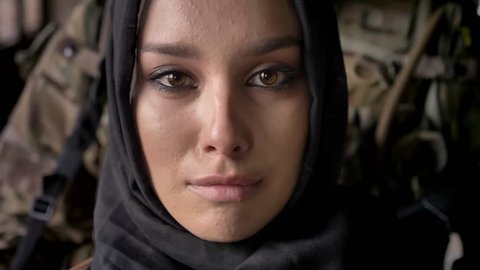 Close portrait of young muslim woman in hijab crying and looking at camera, armed soldier with weapon standing behind woman, war