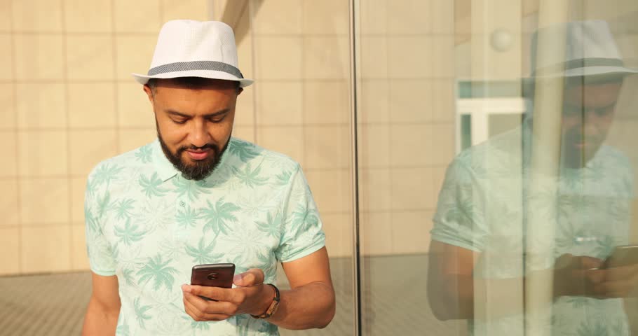People and Technolody. Portrait of the cheerful african man in t-shirt and hat chatting and browsing on the mobile phone, pulling his cuitcase to the aiport. 4k.