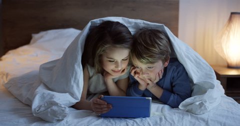 Close up of the cute small boy and girl lying under the blanket at night and watching video on tablet computer. Inside. Social distance, Self isolation, protective, quarantine  epidemic,coronavirus