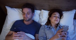 View from above on the Caucasian married man and woman after a quarrel lying in the bed at night and scrolling or texting on the smartphones. Close up. Inside.