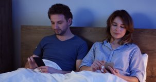 Caucasian married couple of middle age sitting in their bed at night and scrolling or taping on their smartphones. Indoor.