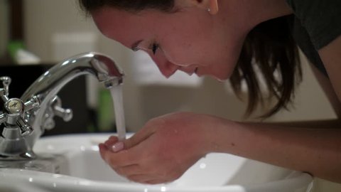 Woman splash water on face, stay over sink at washing room. Young adult lady feel tired at evening, closeup shot. She hold cupped hands then rise up to face