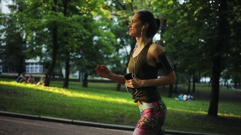 Beautiful runner woman running in park exercising outdoors fitness tracker slow motion sunlight healthy young jogging music sport girl summer nature technology active athlete workout sunset body