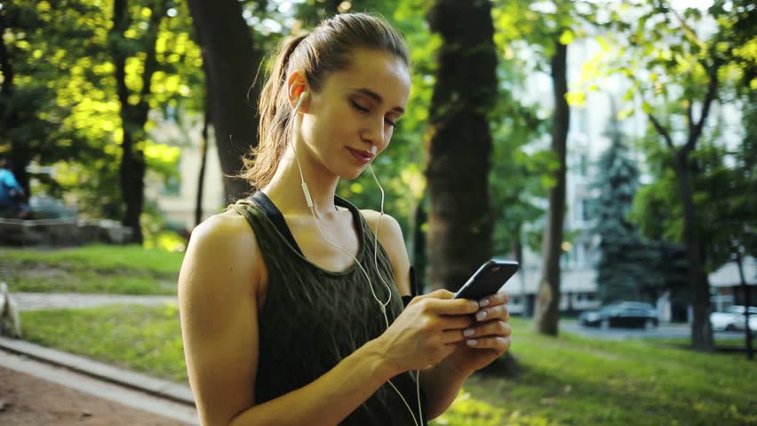 Happy young woman use smartphone and headphone standing in park sun outdoor health hand girl tree sport fitness summer nature internet technology female runner exercise phone workout mobile cellphone Royalty-Free Stock Footage #1013464853