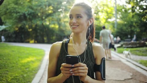 Happy young woman use smartphone and headphone standing in park sun outdoor health hand girl tree sport fitness summer nature internet technology female runner exercise phone workout mobile cellphone