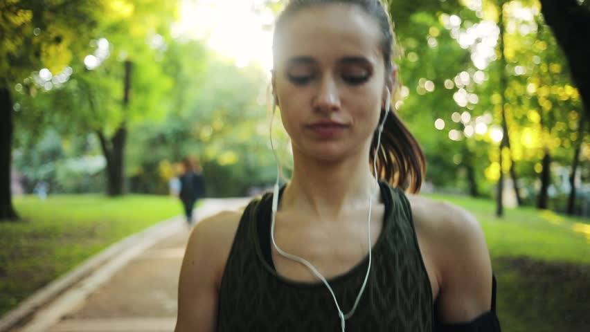 Portrait runner woman running in park use phone exercising outdoors fitness tracker slow motion sunlight healthy young jogging music sport girl summer nature active athlete workout sunset body