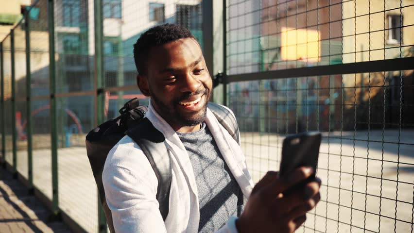 Good-looking student getting mobile phone from pocket. Cute surprised young African man looking at screen of smartphone, laughing. Street. Outdoors.