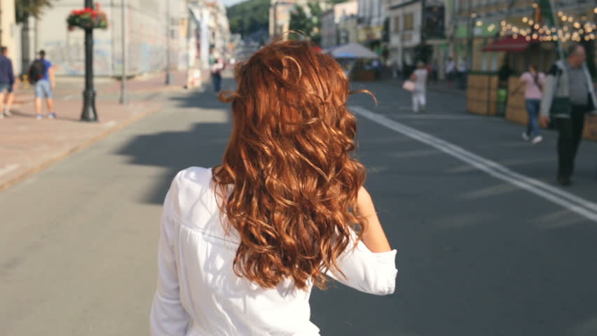 Happy, beatiful curly hair woman listening to music and walking at the streeet. View from back.